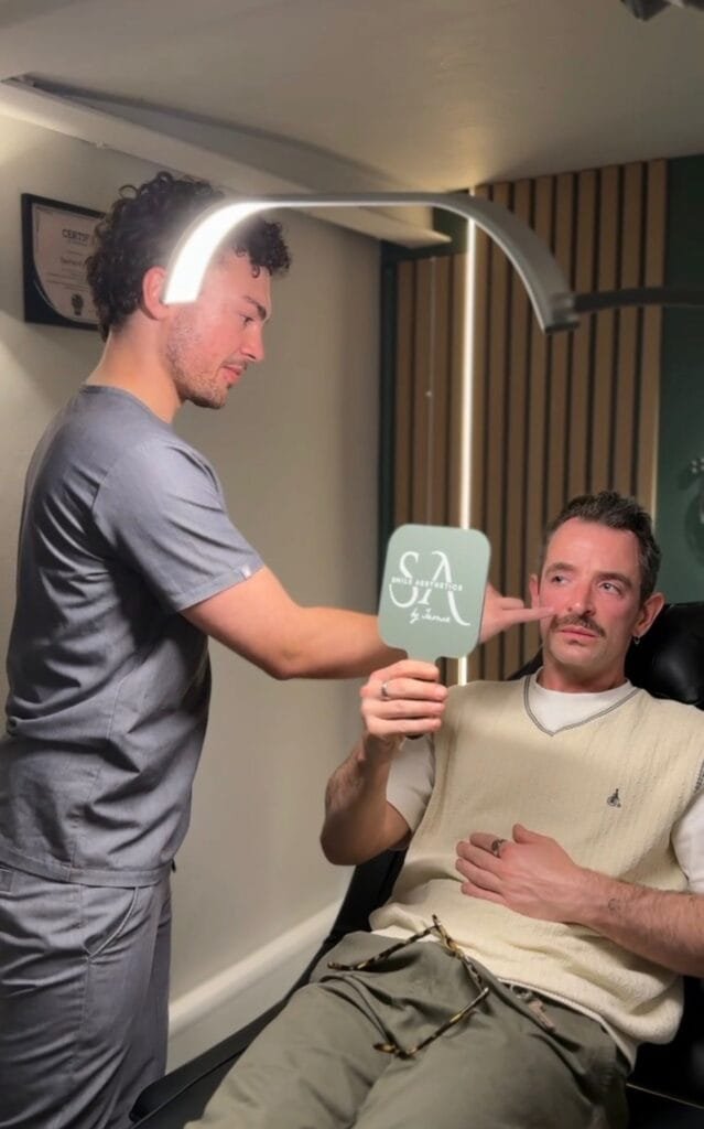 Practitioner Jamie Amphlett holds a mirror for a male patient during a men's aesthetics consultation in Cardiff, as the patient points to his face to discuss natural results.