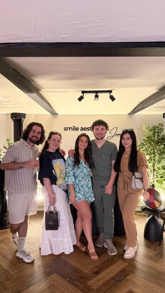A group of five colleagues and clients posing together in the reception area of Smile Aesthetics Clinic, with the clinic's logo visible on the wall behind them. New clinic Cardiff.